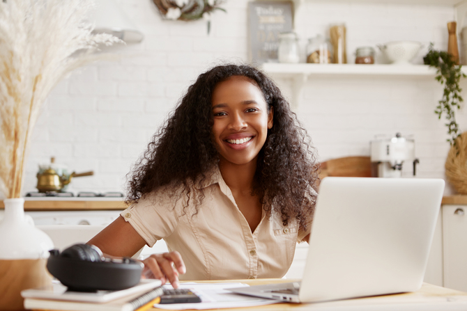 Attractive stylish young dark skinned female in beige shirt sitting at kitchen table, using laptop, calculating budget, planning vacations, smiling happily. Self employed black woman working from home